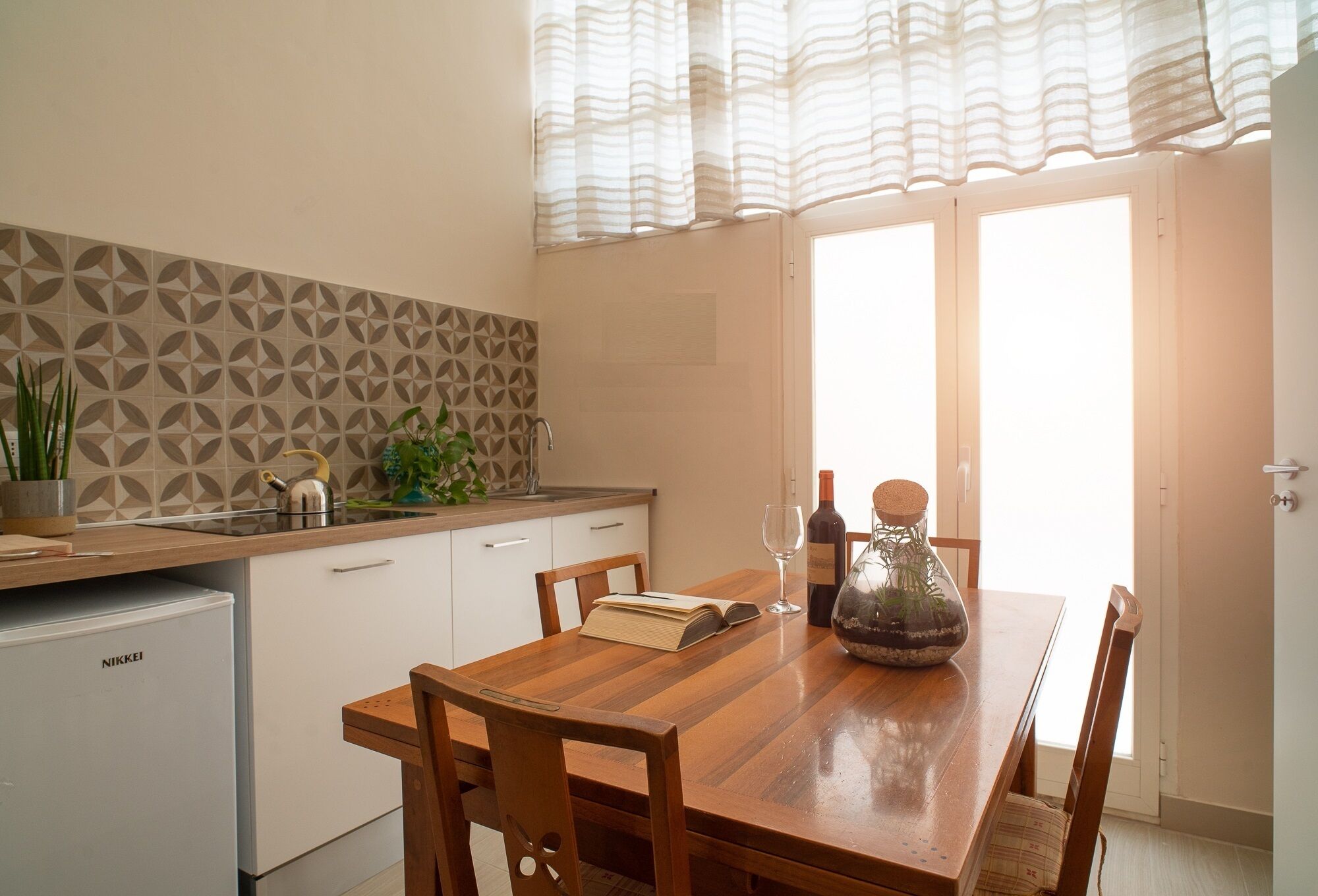 Una cocina con una mesa de madera, sillas, un refrigerador y una ventana con cortinas.