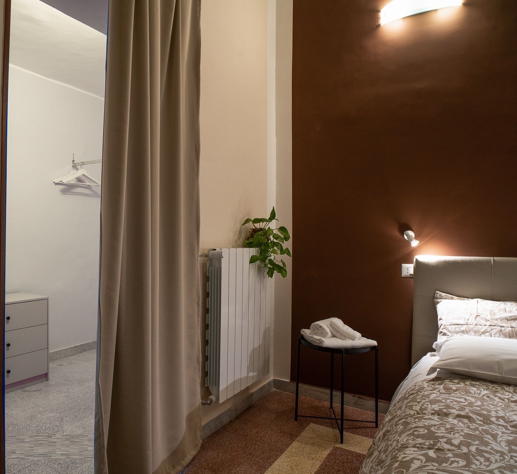 Une chambre avec un lit, un radiateur, une plante suspendue et une serviette posée sur un tabouret.