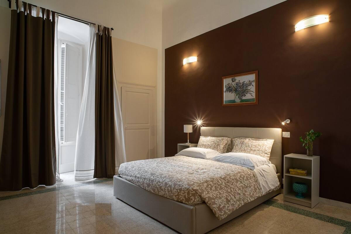 A bedroom with a large bed, two bedside tables, a nightstand, a wall-mounted light, and a door with curtains.