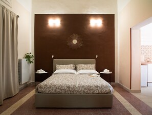 A neatly arranged hotel room with a large bed, two bedside tables, a wall-mounted light fixture, and a radiator. - Corte Nicolaiana Apts by Wonderful Italy (Bari)