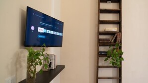 Μια τηλεόραση με επίπεδη οθόνη είναι τοποθετημένη στον τοίχο και στην οθόνη εμφανίζεται η διεπαφή της Netflix. Κάτω από την τηλεόραση υπάρχει ένα μαύρο ράφι με μια γλάστρα και ένα βιβλίο. Στα δεξιά, υπάρχει ένα ξύλινο ράφι σε σχήμα σκάλας, με βιβλία και διακοσμητικά αντικείμενα.