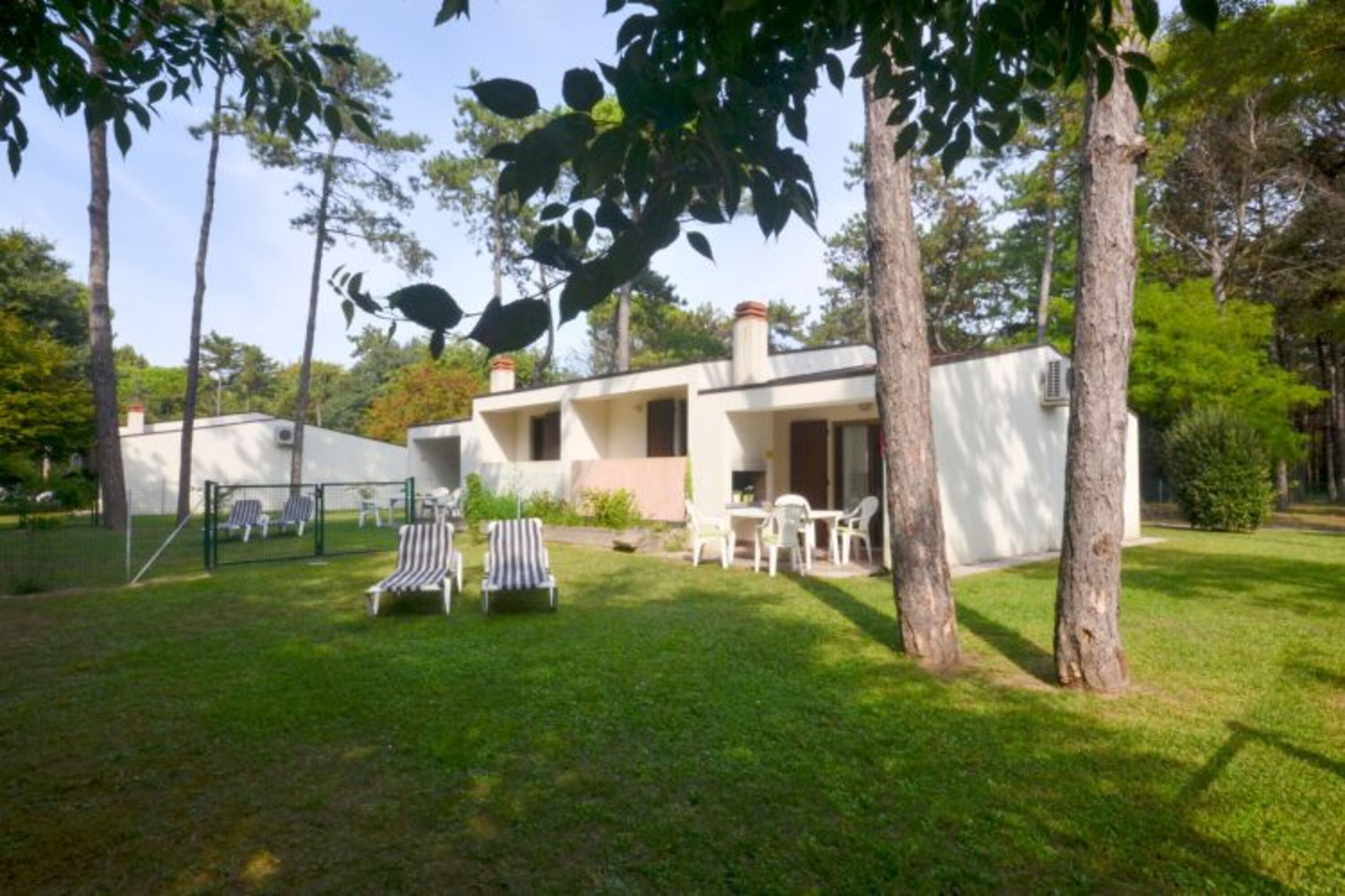 A modern building with a flat roof, surrounded by a well-maintained lawn and trees. There are outdoor seating areas with chairs and tables.