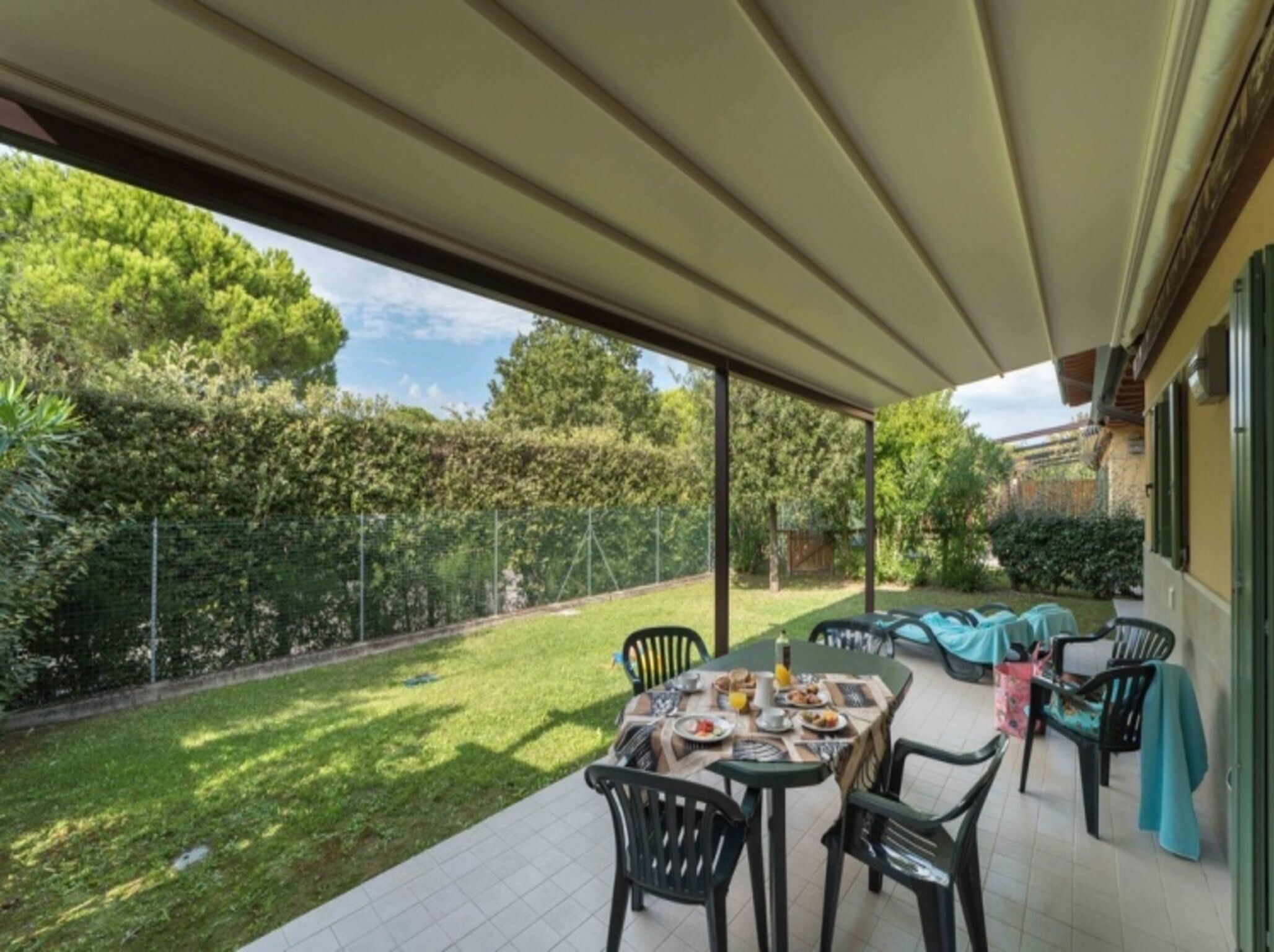 A covered patio with a table set for breakfast, surrounded by chairs, overlooking a well-maintained garden and a fence.