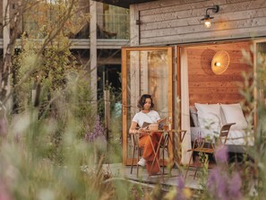 A person sitting at a table outside a wooden cabin with a glass door, surrounded by greenery. - Hôtel Le Bout du Parc (Versailles)