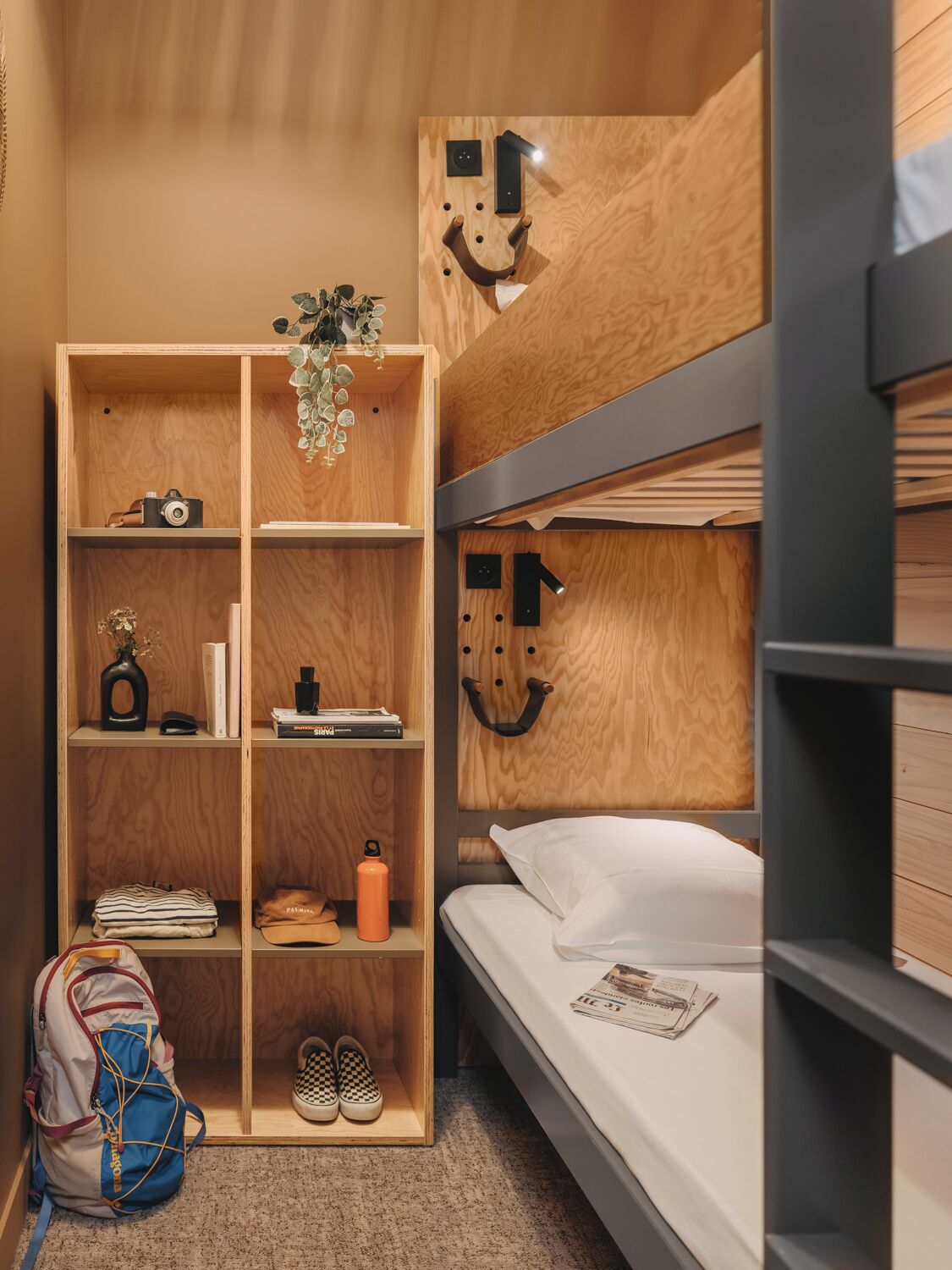 Een slaapkamer met een stapelbed, een houten hoofdbord, een boekenkast en een klein bureau met een camera en boeken.