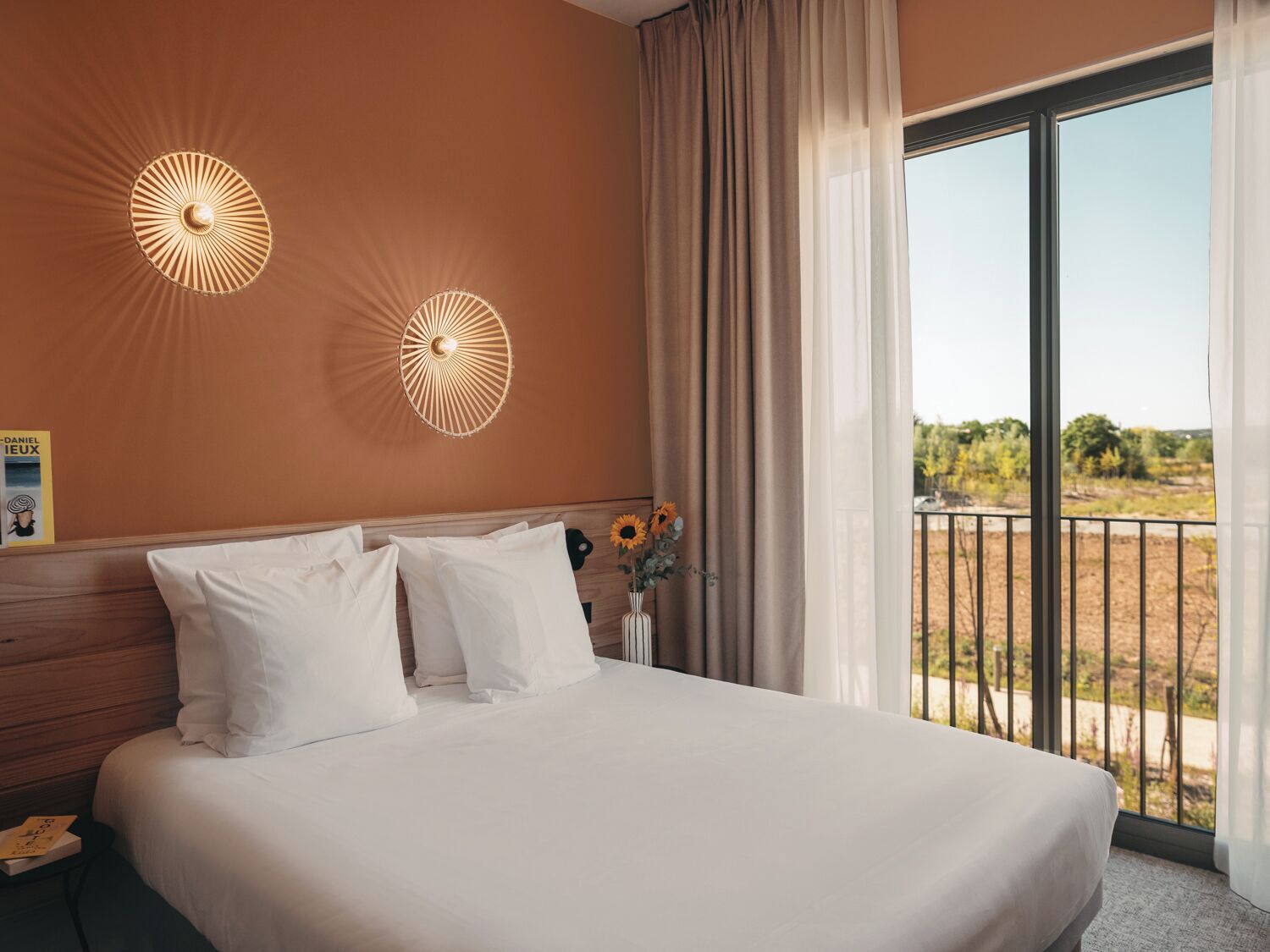 Une chambre d’hôtel avec un grand lit, deux lampes fixées au mur, une fenêtre donnant sur l’extérieur et un vase rempli de fleurs placé sur la table de chevet.