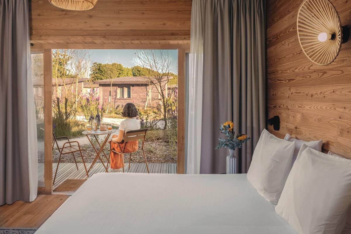 Quarto com uma cama grande, teto de madeira e vista para um jardim e um edifício.