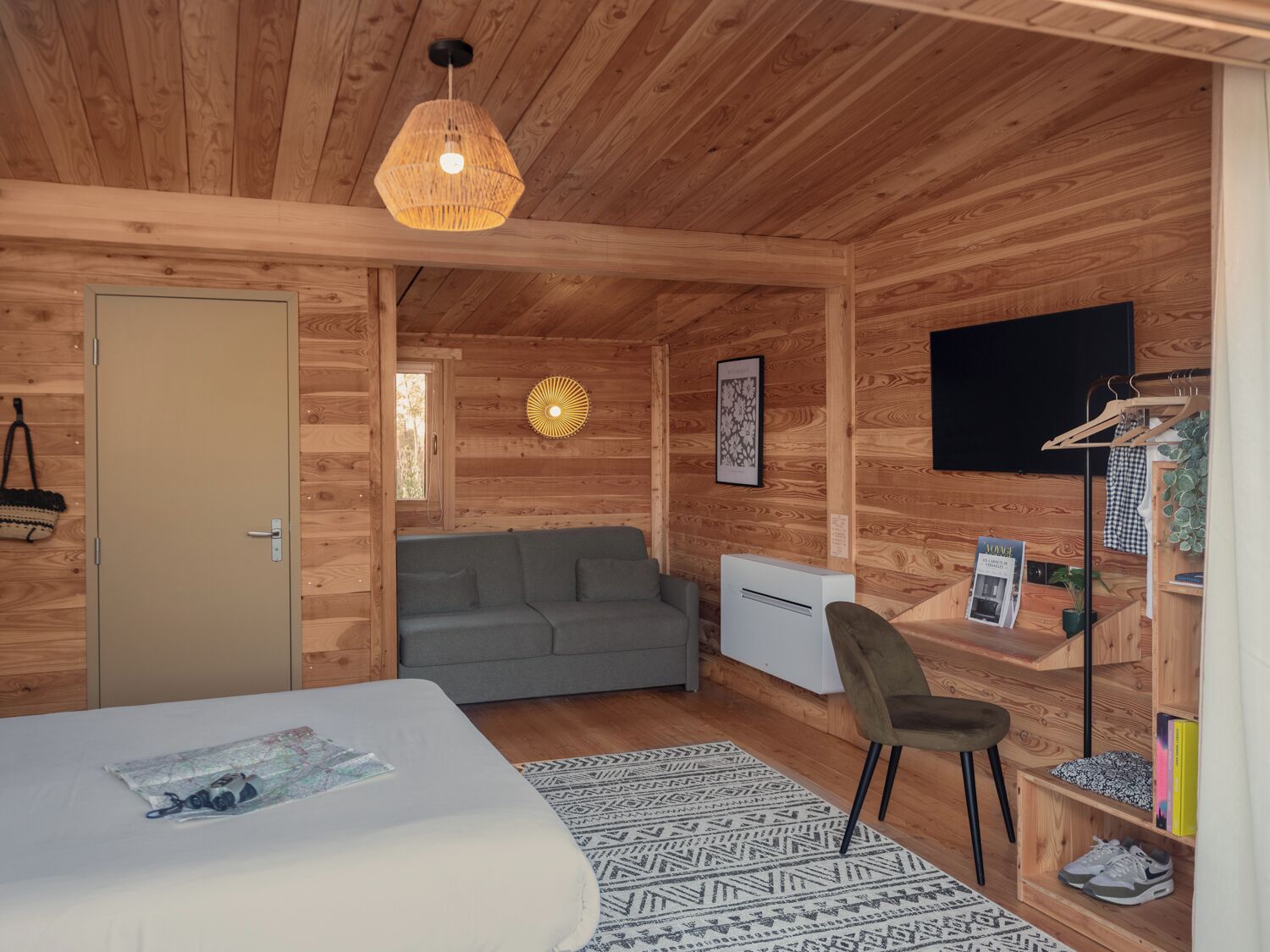 A wooden cabin room with a bed, a grey sofa, a desk with a chair, a TV, and a small plant.