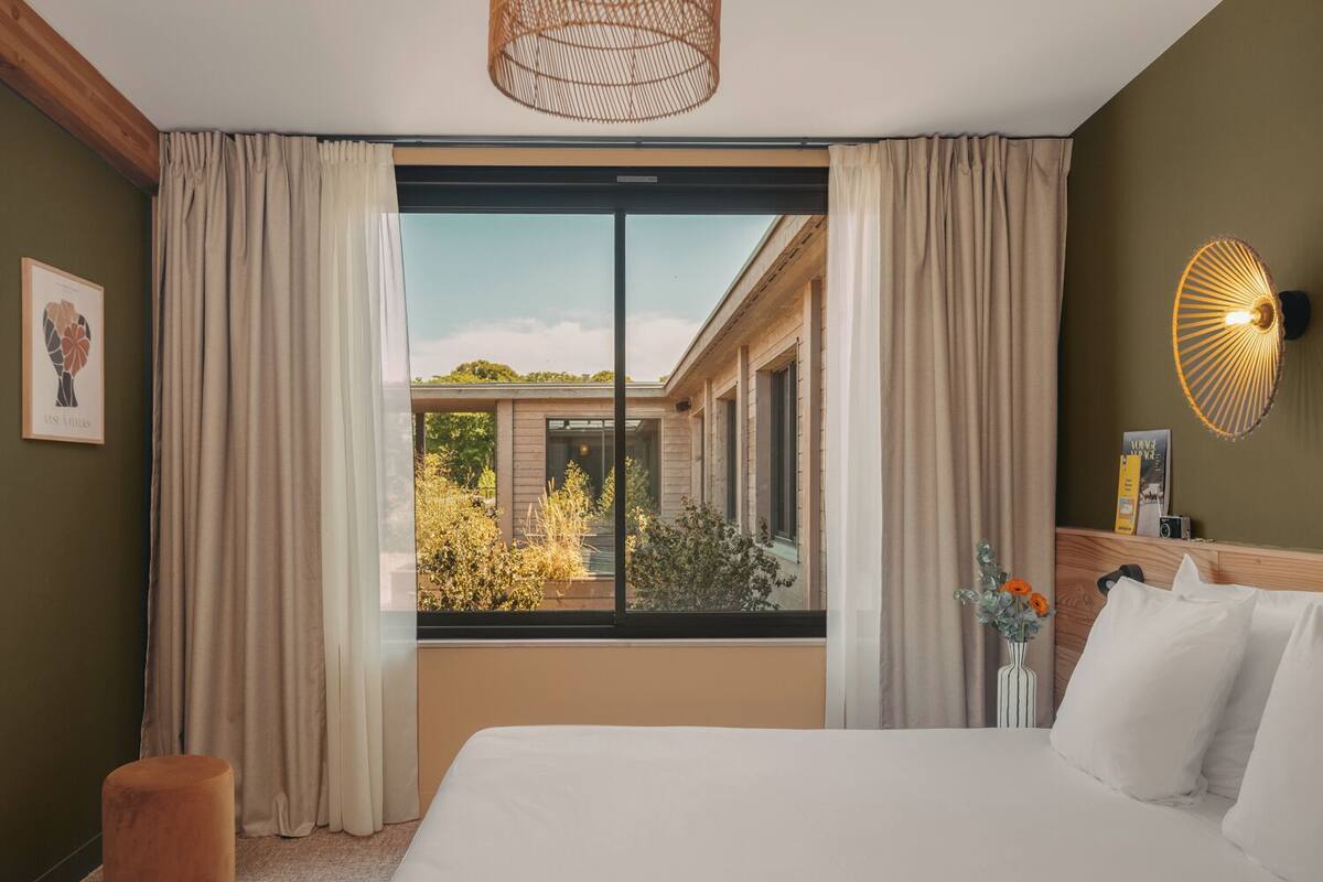 Quarto de hotel moderno com uma janela grande, uma cama, uma mesa de cabeceira e um lustre.