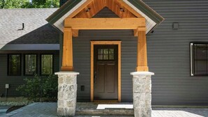 Una casa moderna con fachada gris, un porche delantero de madera sostenido por pilares de piedra y una puerta de color marrón oscuro.