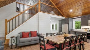 Amplia sala de estar con escalera de madera, un sofá gris con cojines rojos, una mesa de comedor de madera con sillas, y una cocina con armarios grises y una ventana.
