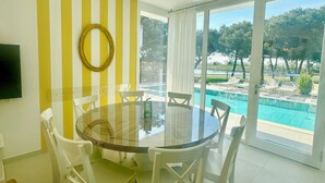 A dining area with a round glass table and white chairs, a yellow-striped wall, and a view of a swimming pool and trees. - Stunning villa with pool and lagoon view (Isola di Albarella)