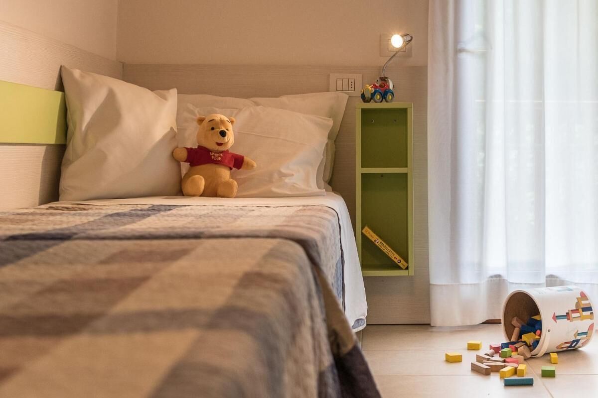 Un dormitorio con una cama, almohadas, un osito de peluche, un coche de juguete y bloques de construcción desperramados.