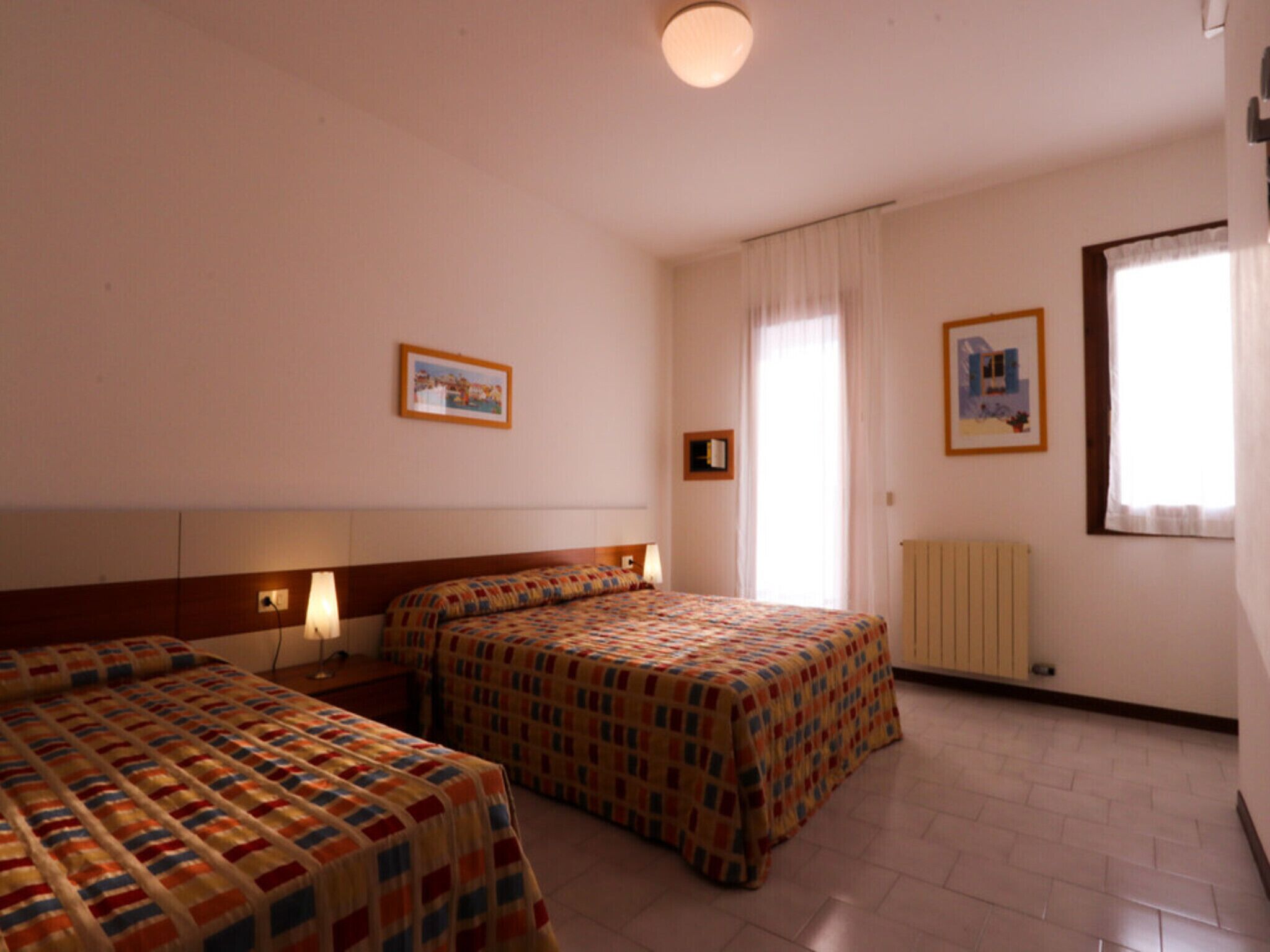 Ein Hotelzimmer mit zwei Betten, einem großen Fenster mit Vorhängen, einem Heizkörper und gerahmten Bildern an den Wänden.