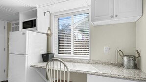Una cocina con armarios blancos, un refrigerador, una ventana con persianas, una silla y una olla de metal.