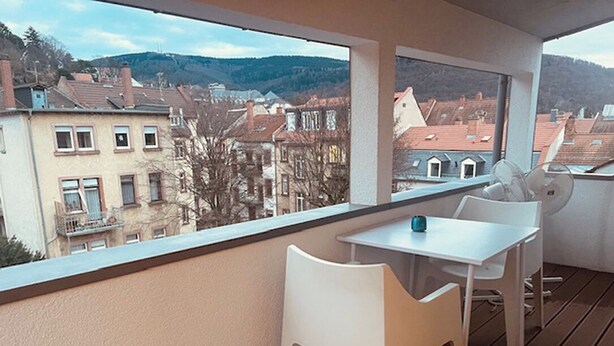 Ein Balkon mit Tisch und zwei Stühlen, mit Blick auf eine Wohngegend mit Häusern und Hügeln.