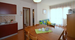 A dining area with a wooden table, chairs, and a green placemat. There is a kitchen area with cabinets and a sink. A window with curtains is visible in the background.