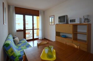 A room with a sofa, a dining table with chairs, a TV, a coffee maker, and a window with a view of buildings.