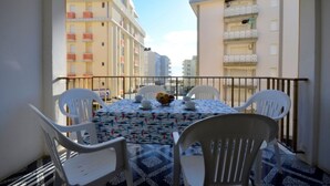 Un balcon avec une table dressée pour deux personnes, des chaises en plastique blanches, un saladier rempli de fruits et une nappe à motifs.