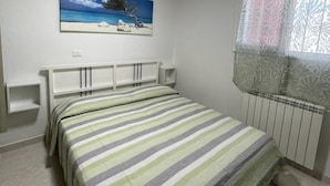 A bedroom with a bed, a painting of a beach scene, a window with patterned curtains, and a radiator. - Spacious apartment with terrace in Rosolina Mare (Rosolina Mare)