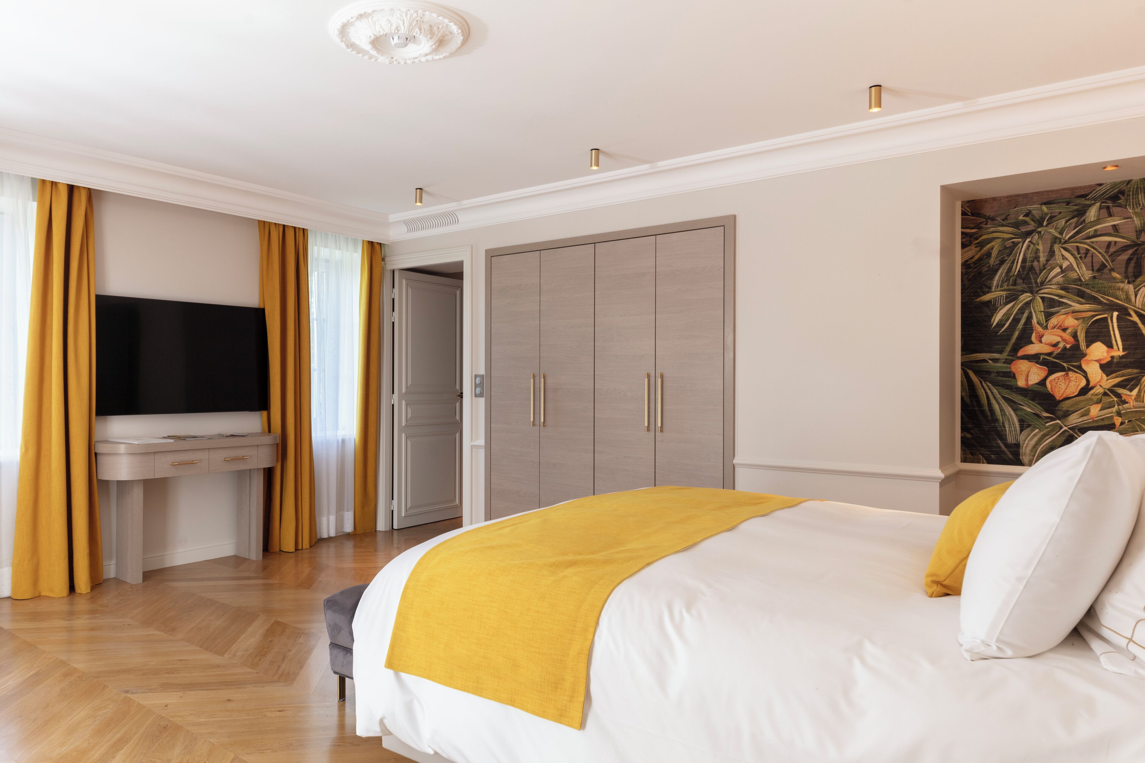 A hotel room with a bed, a desk with a flat-screen TV, a wardrobe, and a painting on the wall.