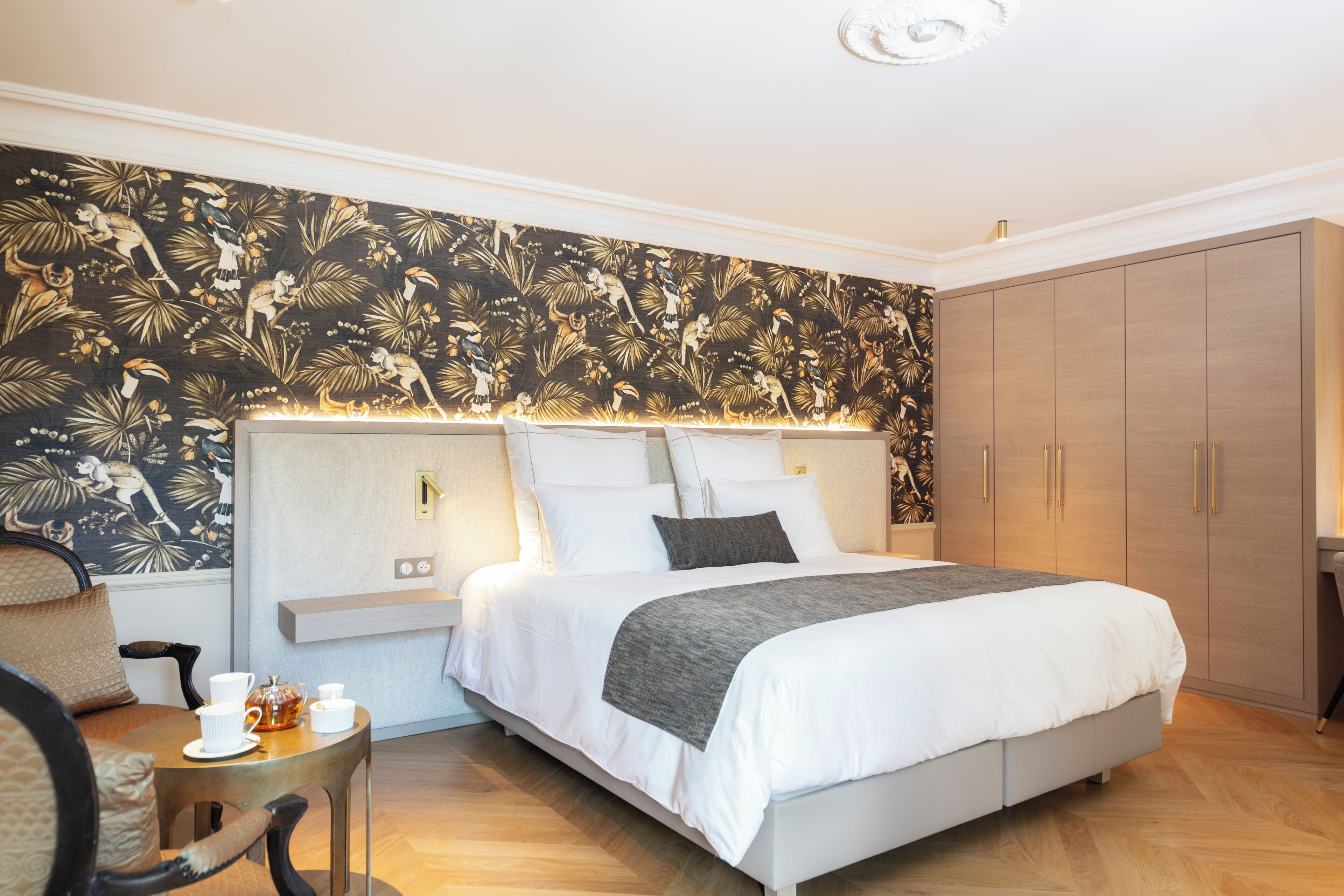 A modern hotel room with a large bed, a nightstand, a small table with a cup and a teapot, and a wardrobe.