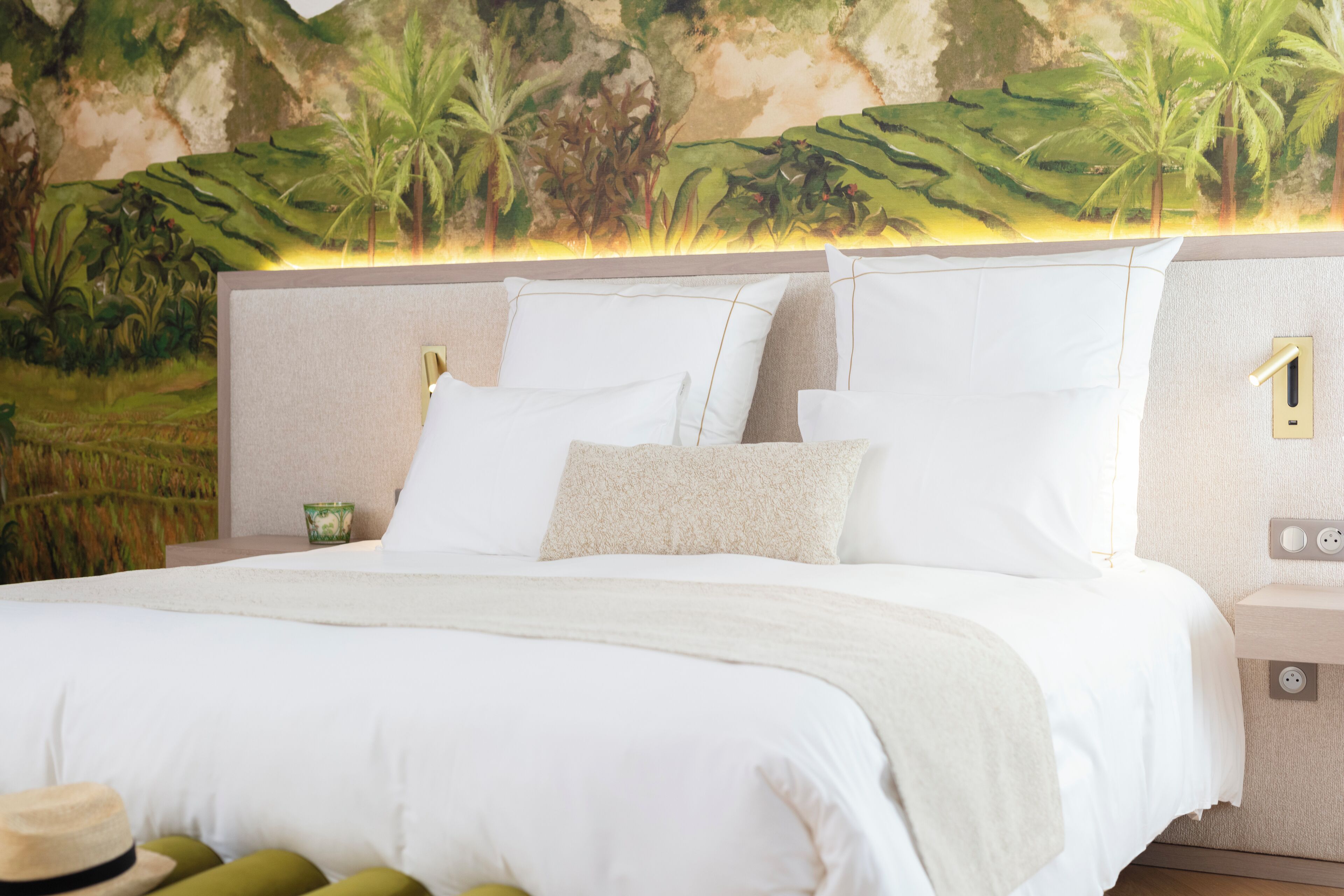 Un lit bien fait, avec du linge de lit blanc et un plaid beige, une table de chevet avec une lampe verte, et une fresque murale représentant un paysage tropical.