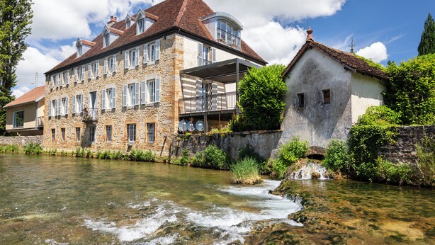 A stone building with a river in front, featuring a balcony and a smaller adjacent building. - Le Clos Alice (Arbois)