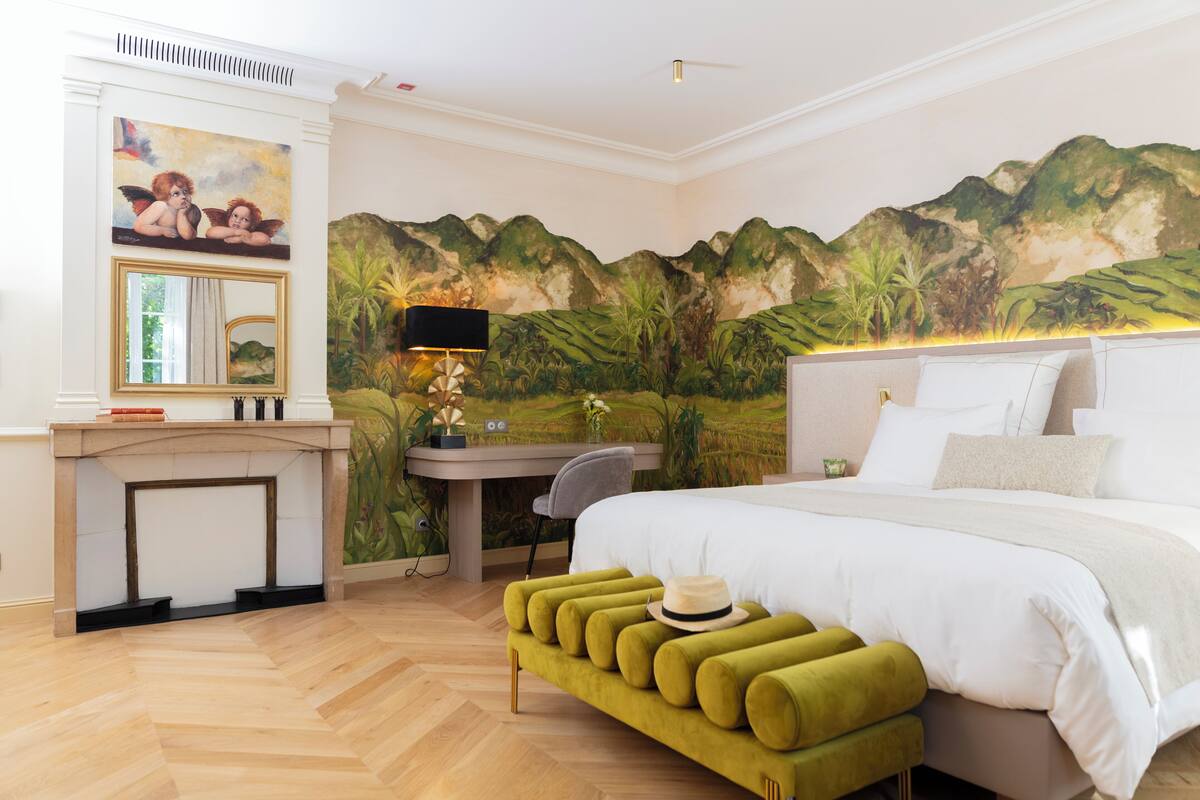 A bedroom with a bed, a desk, a chair, a green bench, a mirror, and a mural on the wall.