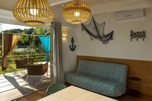 A room with a wooden table, a patterned sofa, wicker chairs, and a view of greenery outside.