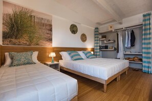 Une chambre avec deux lits, une tête de lit en bois, une table de chevet avec une lampe, une étagère murale avec des vêtements et un grand tableau représentant une scène de plage.