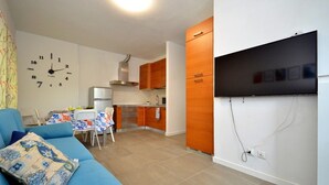 Ett kompakt bostadsutrymme med ett litet kök, en soffa med en dekorativ kudde, en väggklocka och en platt-TV som är monterad på väggen.
