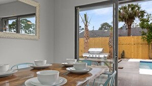 Un comedor con una mesa de madera dispuesta para cuatro, un espejo grande en la pared y vistas a un patio trasero con piscina y parrilla.