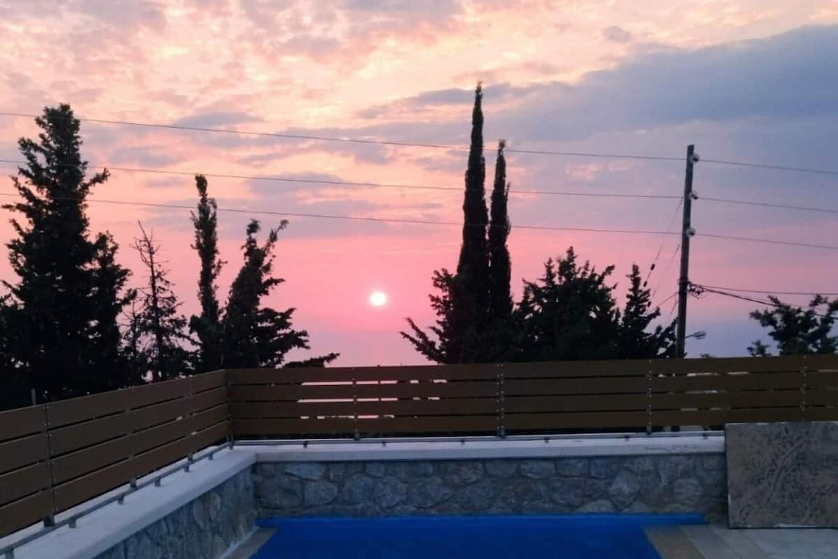 A swimming pool with a stone wall and wooden fence, surrounded by trees and a sunset in the background.