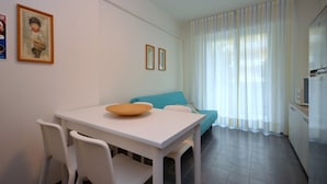 Une petite cuisine moderne avec des meubles blancs, une table à manger avec des chaises, un canapé bleu et une grande fenêtre ornée de rideaux légers.
