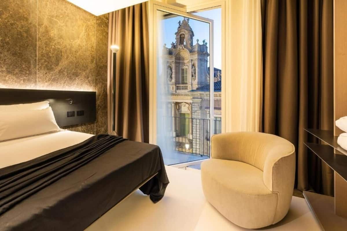 A hotel room with a large bed, a chair, and a view of a historic building through a glass door.
