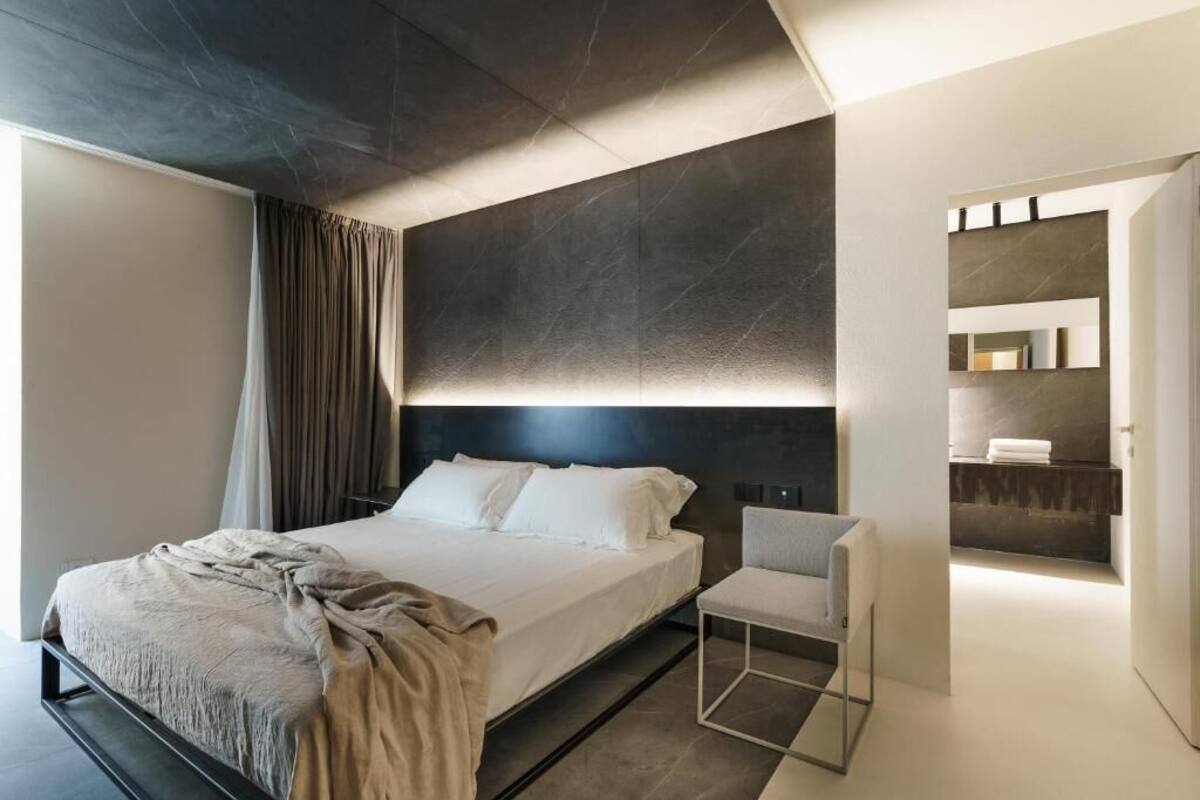 A modern bedroom with a large bed, a bedside chair, a dark headboard, and a built-in shelf with towels.