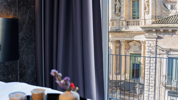 A hotel room with a bed, a tray of pastries, a vase with flowers, and a view of a historic building. - Meo Design Suite & Spa (Catania)
