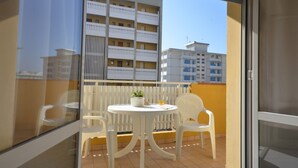Un balcón con una mesa y sillas blancas, una planta en maceta y vistas a un edificio con balcones.