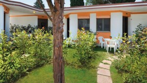 Ein Gebäude mit weißer Fassade und orangen Akzenten, umgeben von einem gepflegten Garten mit einem Baum im Zentrum, einem gepflasterten Weg und Sitzmöglichkeiten im Freien.