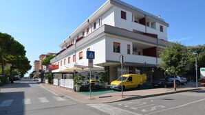 Un immeuble à plusieurs étages avec une façade blanche, un panneau indiquant un passage piéton et un fourgon jaune garé devant.