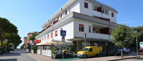 Un edificio de varios pisos con fachada blanca, una señal de paso para peatones y una furgoneta amarilla estacionada adelante.