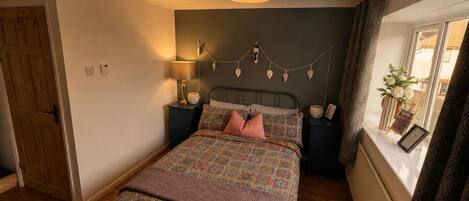 A bedroom with a patterned bedspread, a bedside lamp, a wall-mounted light, a window with curtains, and a wooden door.