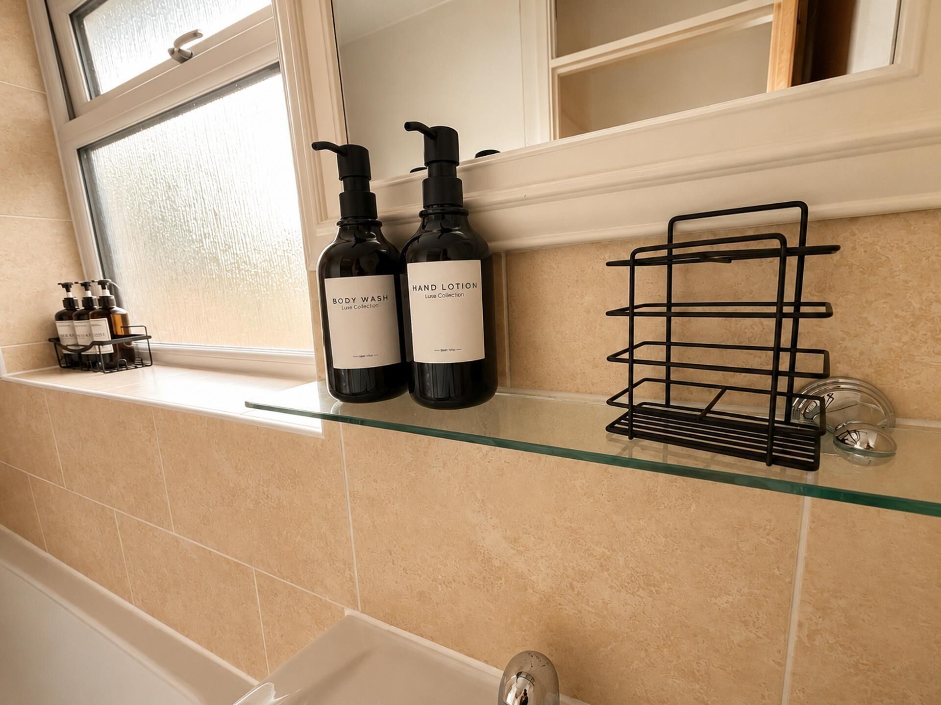 A bathroom shelf with two bottles of body wash and hand lotion, a black soap dispenser, and a glass shelf with a metal rack.