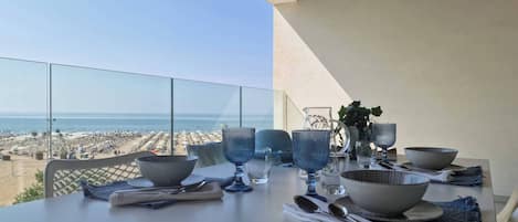 A dining table set with cutlery, bowls, and glasses, overlooking a beach with umbrellas and people.