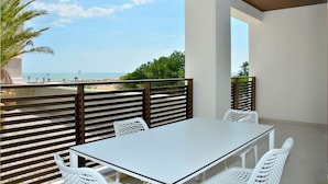 Un balcón con mesa y sillas blancas, vista al mar y una palmera.