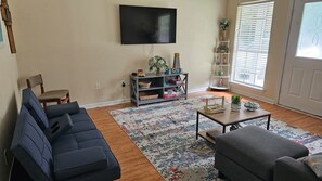 Ein Wohnzimmer mit einer Couch, einem Couchtisch, einem Fernseher, einem Bücherregal und einem Fenster mit Jalousien.