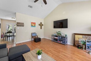 A living room with a flat-screen TV, a dining area with a table and chairs, and a cross on the wall.