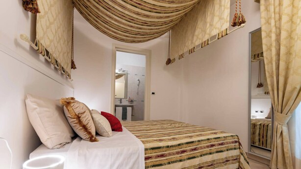 Un dormitorio con cama, almohadas, una mesita de noche, una lámpara colgante y un baño visible a través de una puerta abierta.