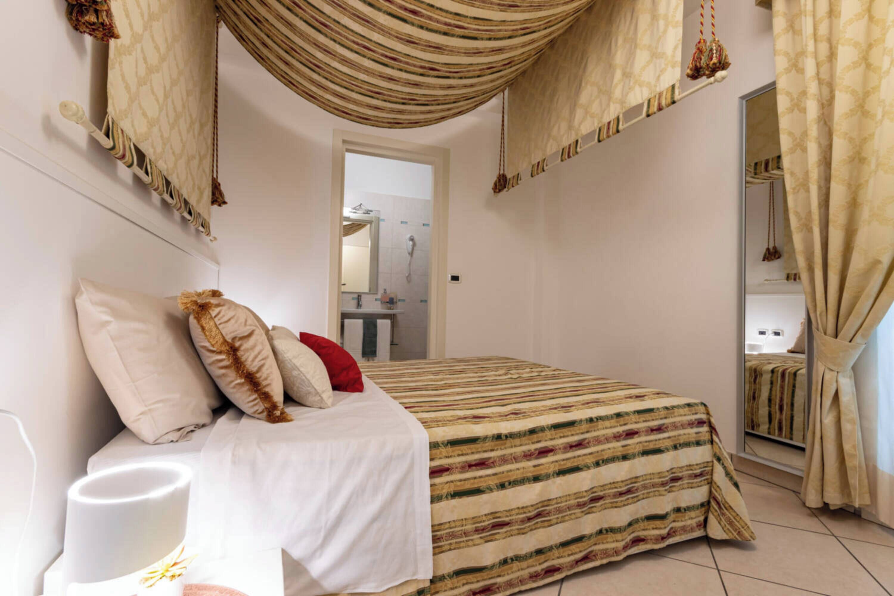 Un dormitorio con cama, almohadas, una mesita de noche, una lámpara colgante y un baño visible a través de una puerta abierta.